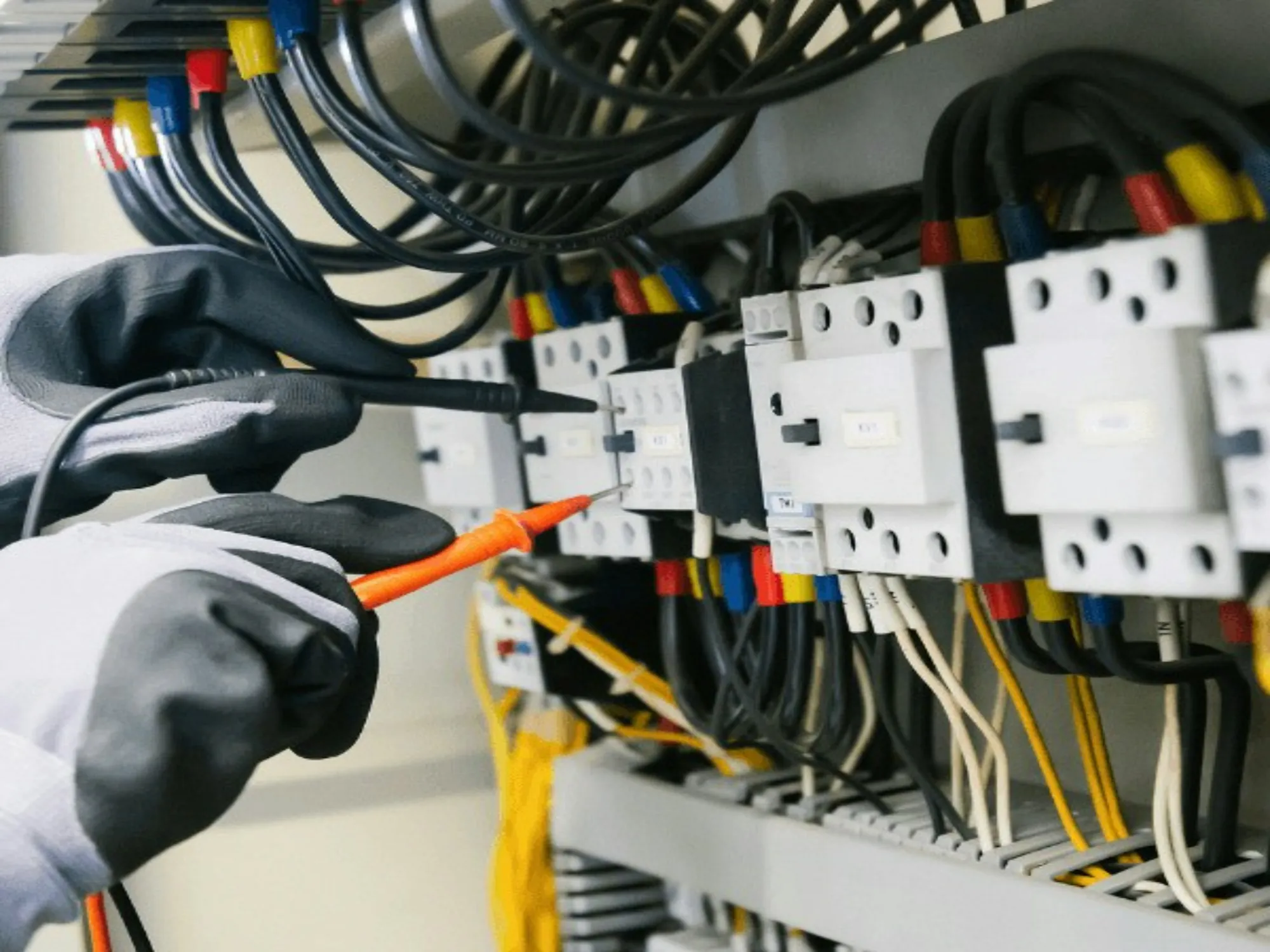 Gloved hands probing wiring on a row of contactors with a voltage tester